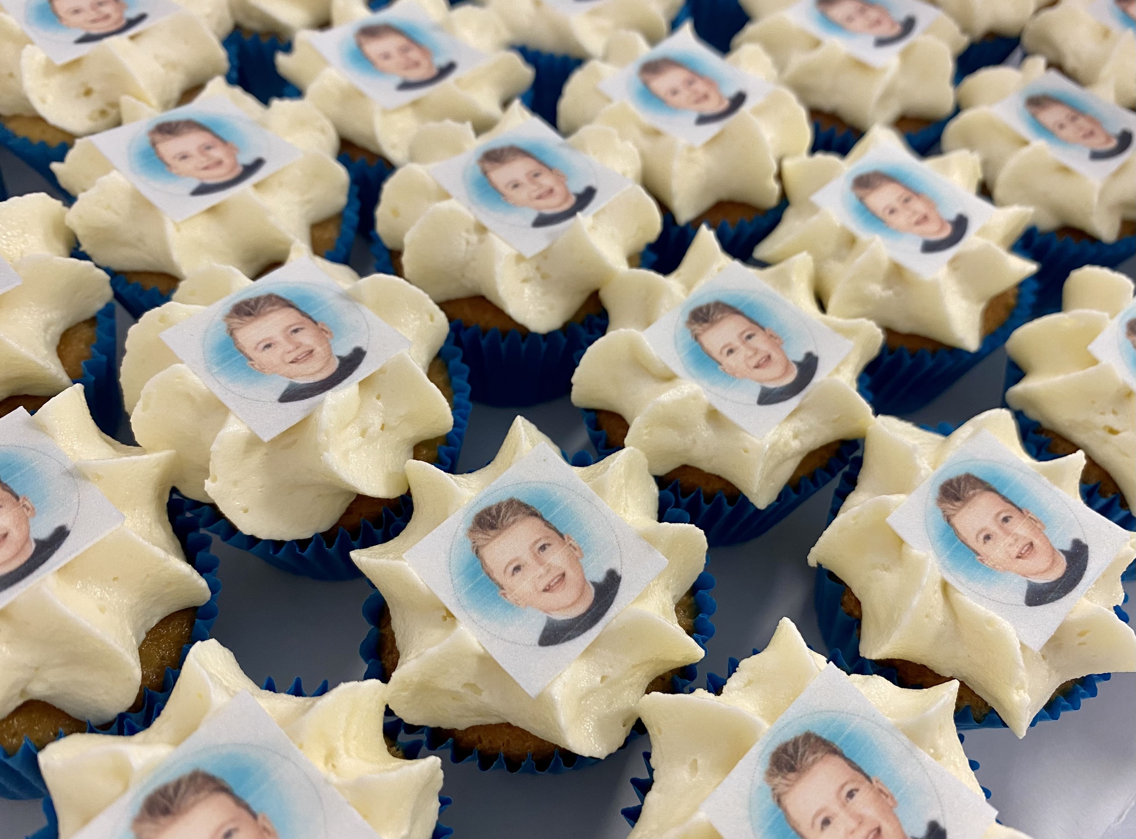 A close-up of numerous custom-designed Mini Vanilla Cupcakes, each topped with white icing and a small, square, edible photo of a young boy with short hair. The cupcakes are arranged in a grid-like pattern on a flat surface.